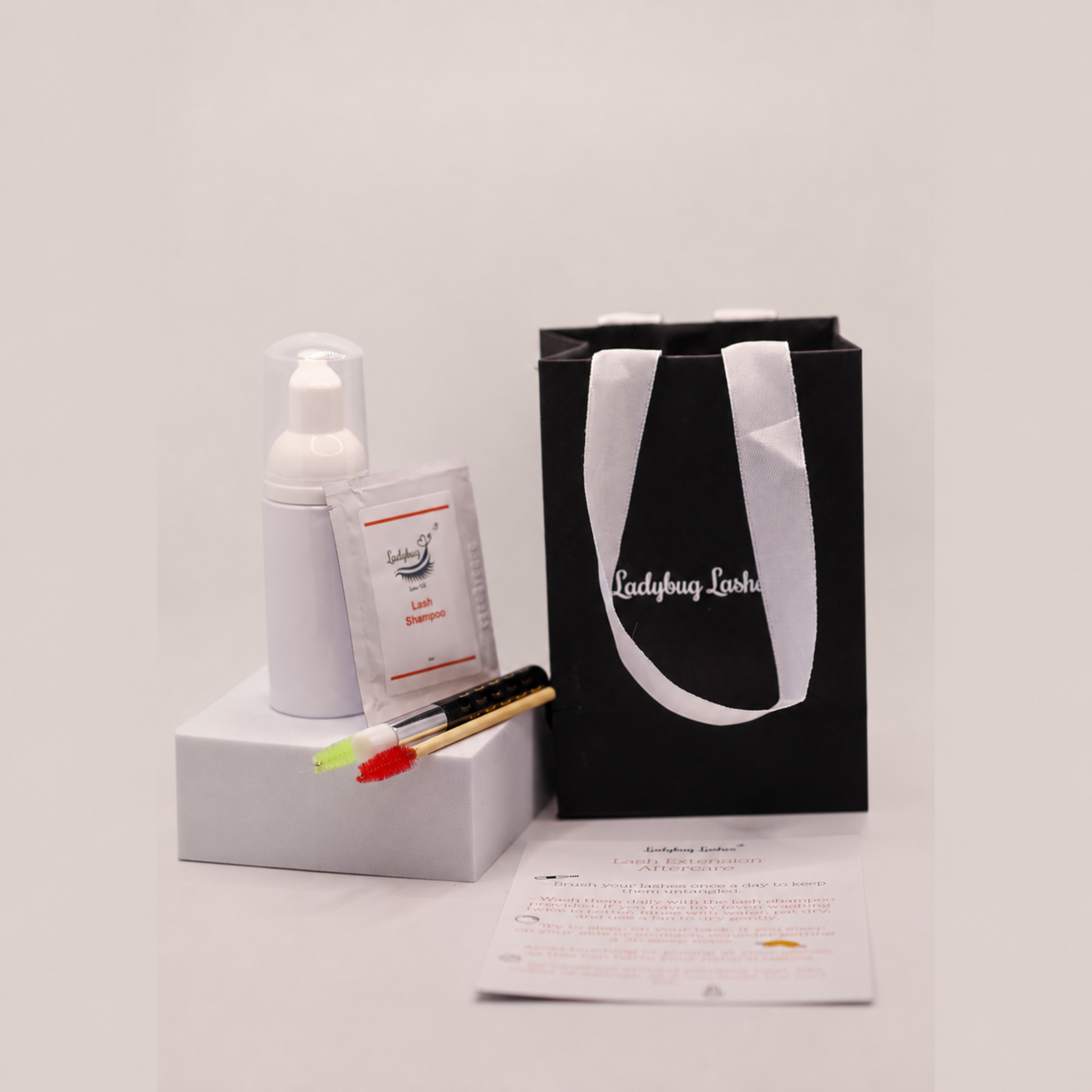 Ladybug Lashes Lash Shampoo Sachet Kit contents displayed on a reflective glass surface surrounded by black Ladybug Lashes gift bags with white satin handles, including a white foam pump bottle, a lash shampoo sachet, a bamboo spoolie, a bamboo cleansing brush with macramé loop, a mini applicator, and a printed Lash Extension Aftercare card