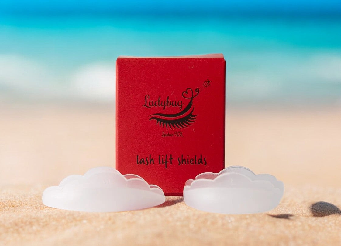 A pair of clear silicone lash lift shields next to a red box with 'LADYBUG LASHES' branding on it, placed on a sandy beach background.
