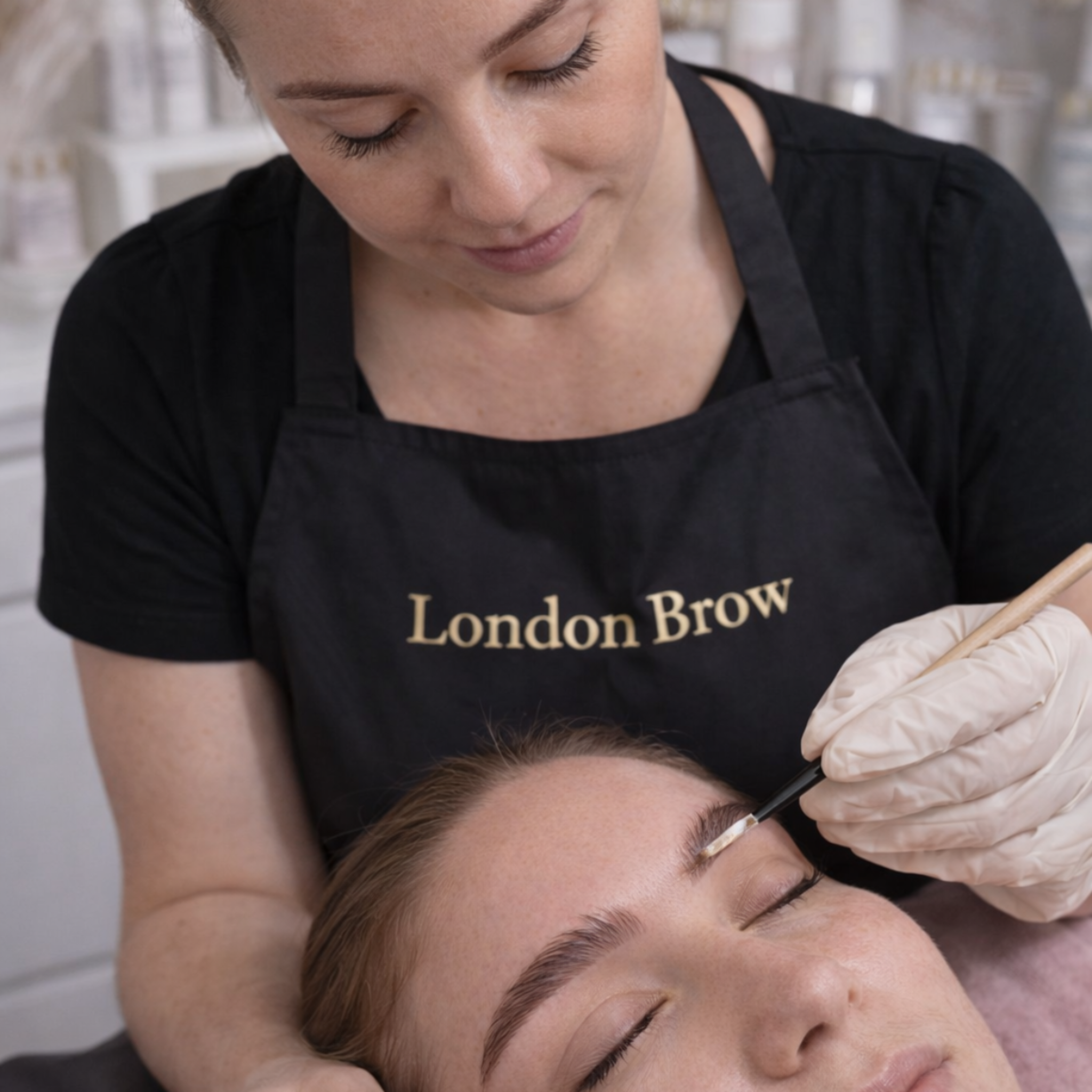 London Brow Company Eyelash and Eyebrow Tinting Course in-treatment shot showing London Brow branded therapist in black apron and latex gloves applying tint to the brow of a fair-skinned female client lying down with eyes closed using a fine precision brush from diagonal overhead angle in a bright salon setting