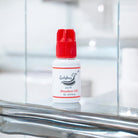 A bottle of Meadow UK 1-2 Second adhesive/glue with a red cap on a reflective surface, branded for Ladybug Lashes.