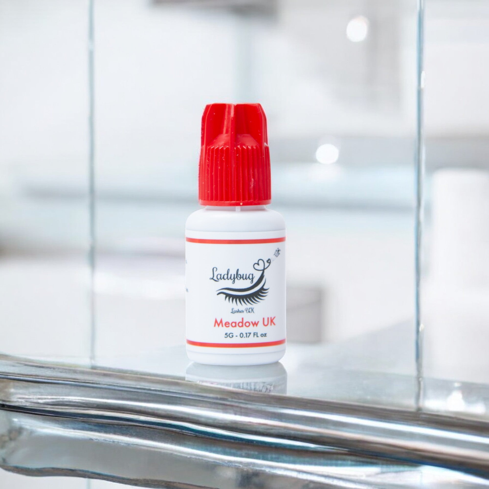 A bottle of Meadow UK 1-2 Second adhesive/glue with a red cap on a reflective surface, branded for Ladybug Lashes.