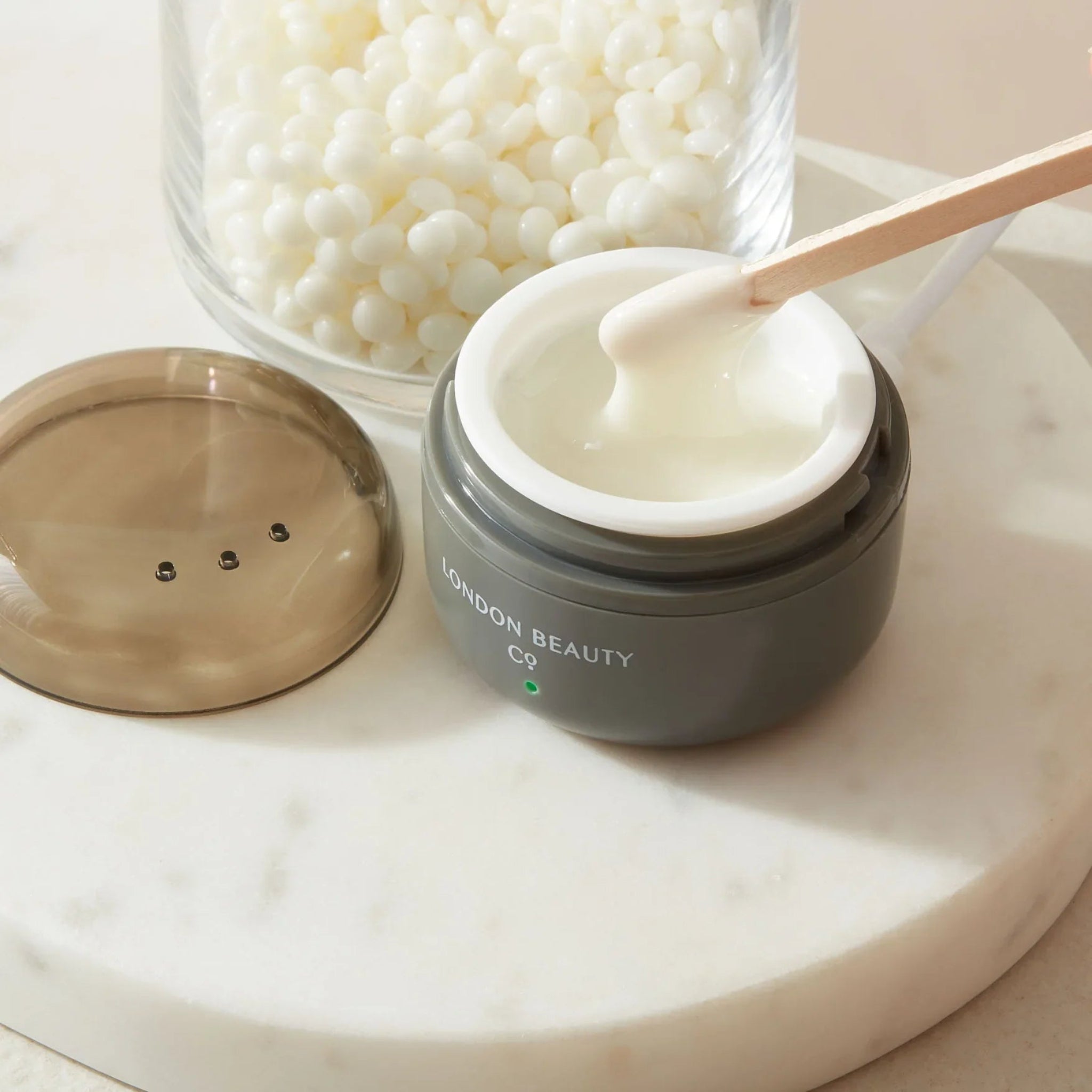 London Beauty Co Mini Portable Wax Heater close-up in-use shot showing dark grey USB-powered mini wax heater with green indicator light open with melted white wax and a wooden spatula lifting wax alongside its brown dome lid and a glass jar of white wax pellets on a white marble tray from diagonal angle