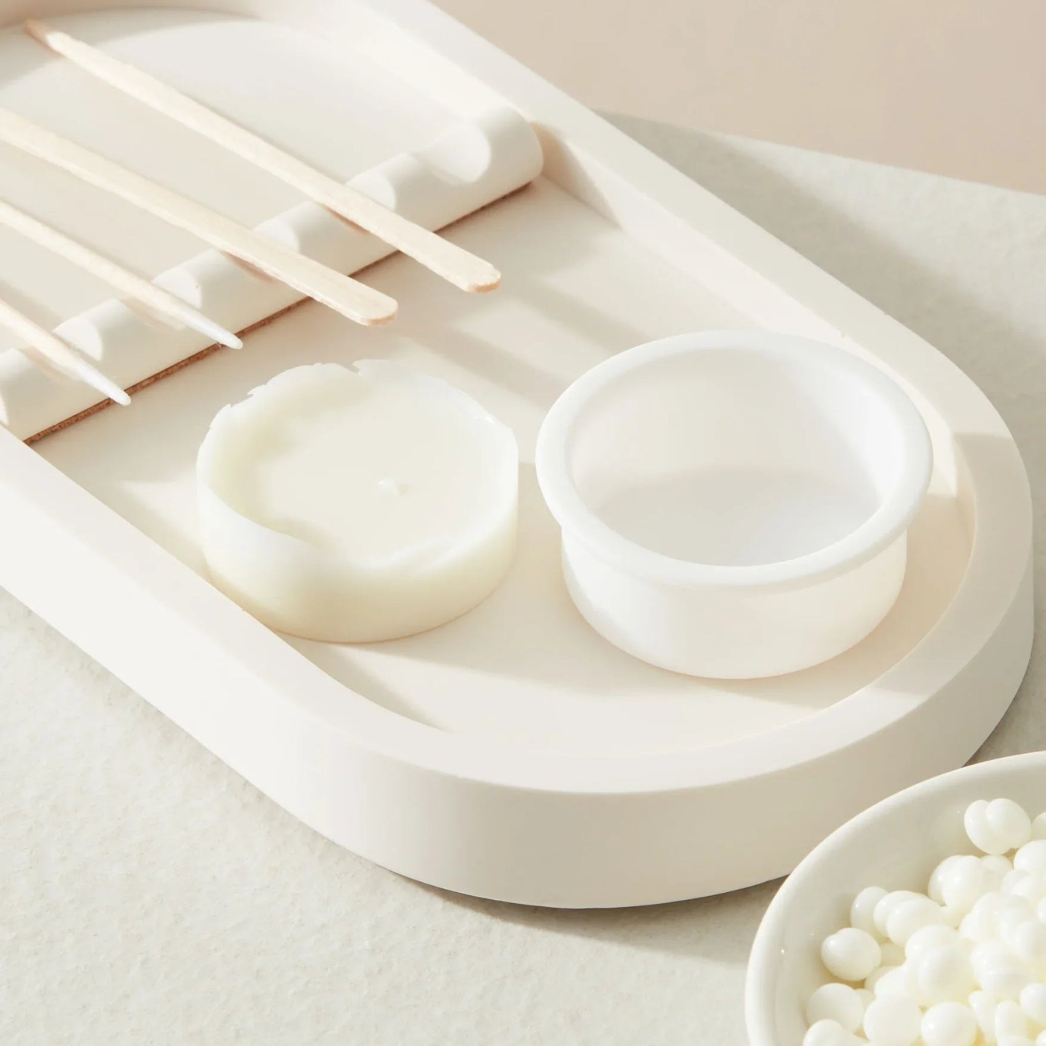 London Beauty Co Mini Portable Wax Heater accessory detail shot showing two white silicone wax pot inserts — one used with set wax and one empty — alongside wooden spatulas and cotton bud applicators on a white oval tray with a bowl of white wax pellets in the foreground from diagonal overhead angle on cream background