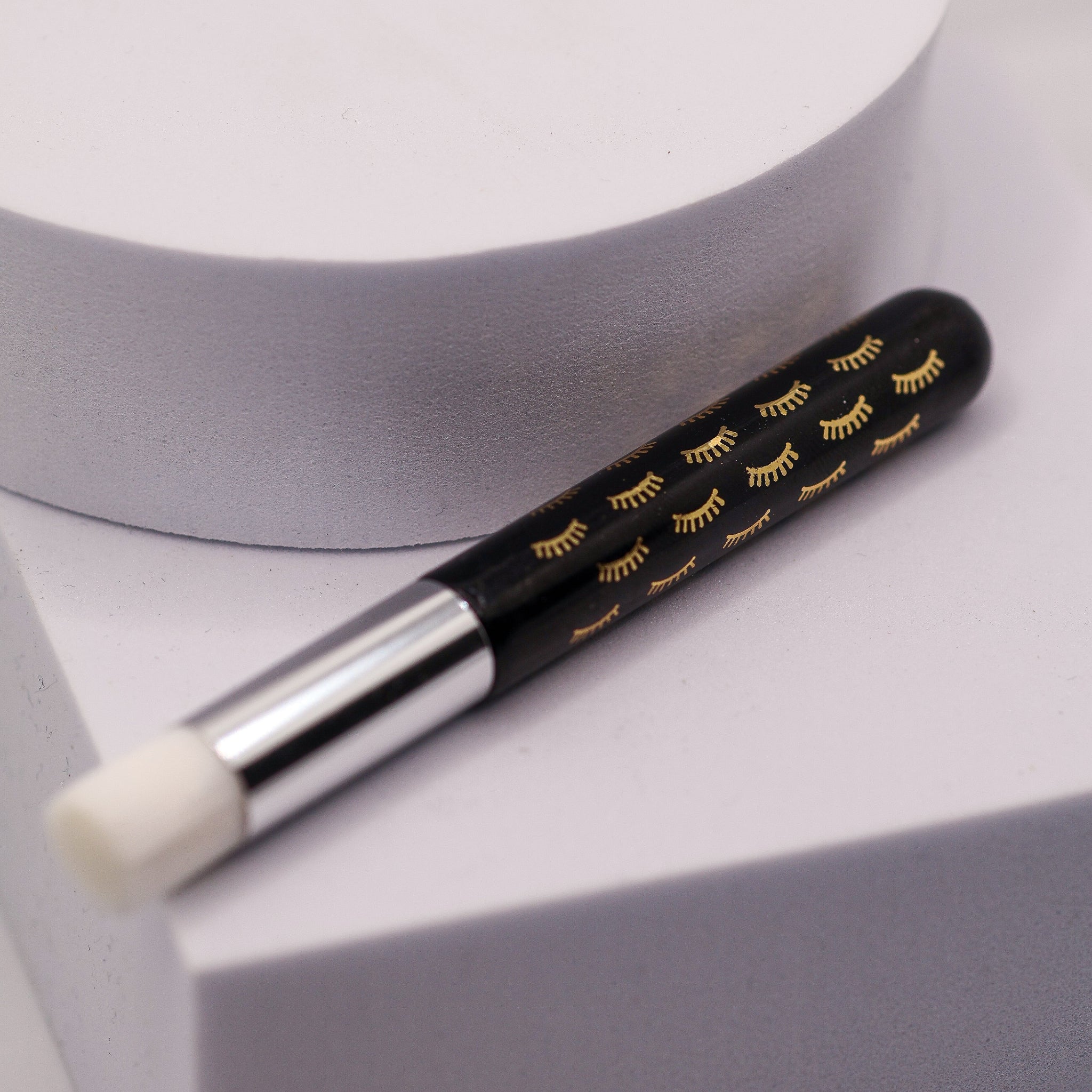 Ladybug Lashes Lash and Brow Cleansing Shampoo Brush with a black handle printed with a repeating gold closed-eye lash pattern and a silver ferrule, lying diagonally on a white cube platform with a round white foam brush head visible, on a light grey background
