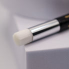 Extreme close-up of the round white foam brush head and silver ferrule of the Ladybug Lashes Lash and Brow Cleansing Shampoo Brush, resting on the edge of a white cube platform against a light grey background, showing the soft dense foam tip used to apply lash shampoo