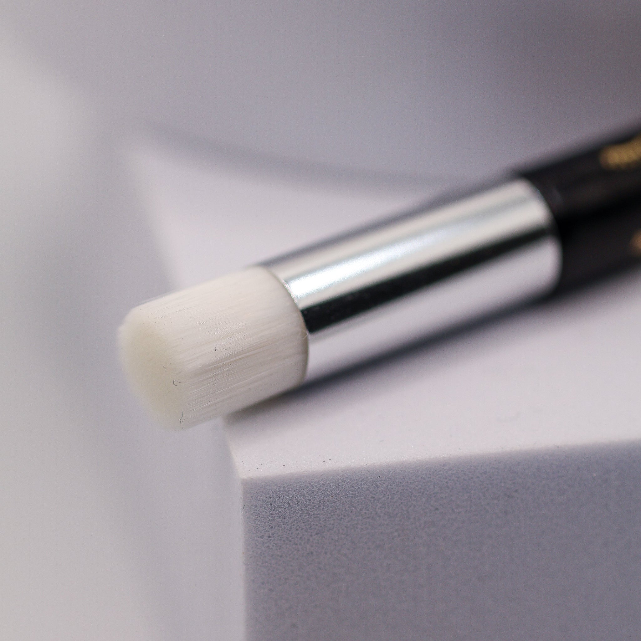 Extreme close-up of the round white foam brush head and silver ferrule of the Ladybug Lashes Lash and Brow Cleansing Shampoo Brush, resting on the edge of a white cube platform against a light grey background, showing the soft dense foam tip used to apply lash shampoo