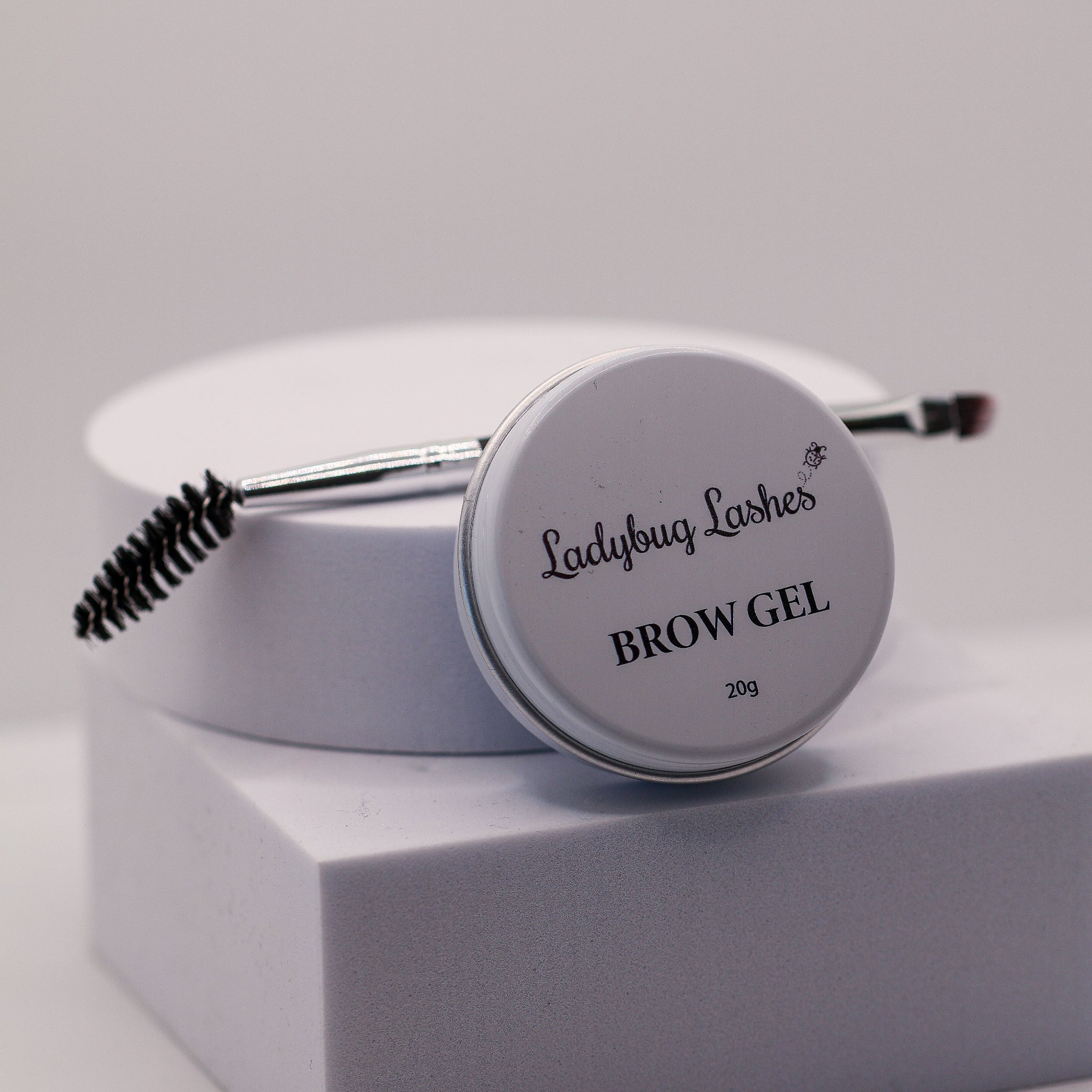 Ladybug Lashes Brow Gel in a round silver tin, displayed on a light gray pedestal with its spoolie brush resting on top.