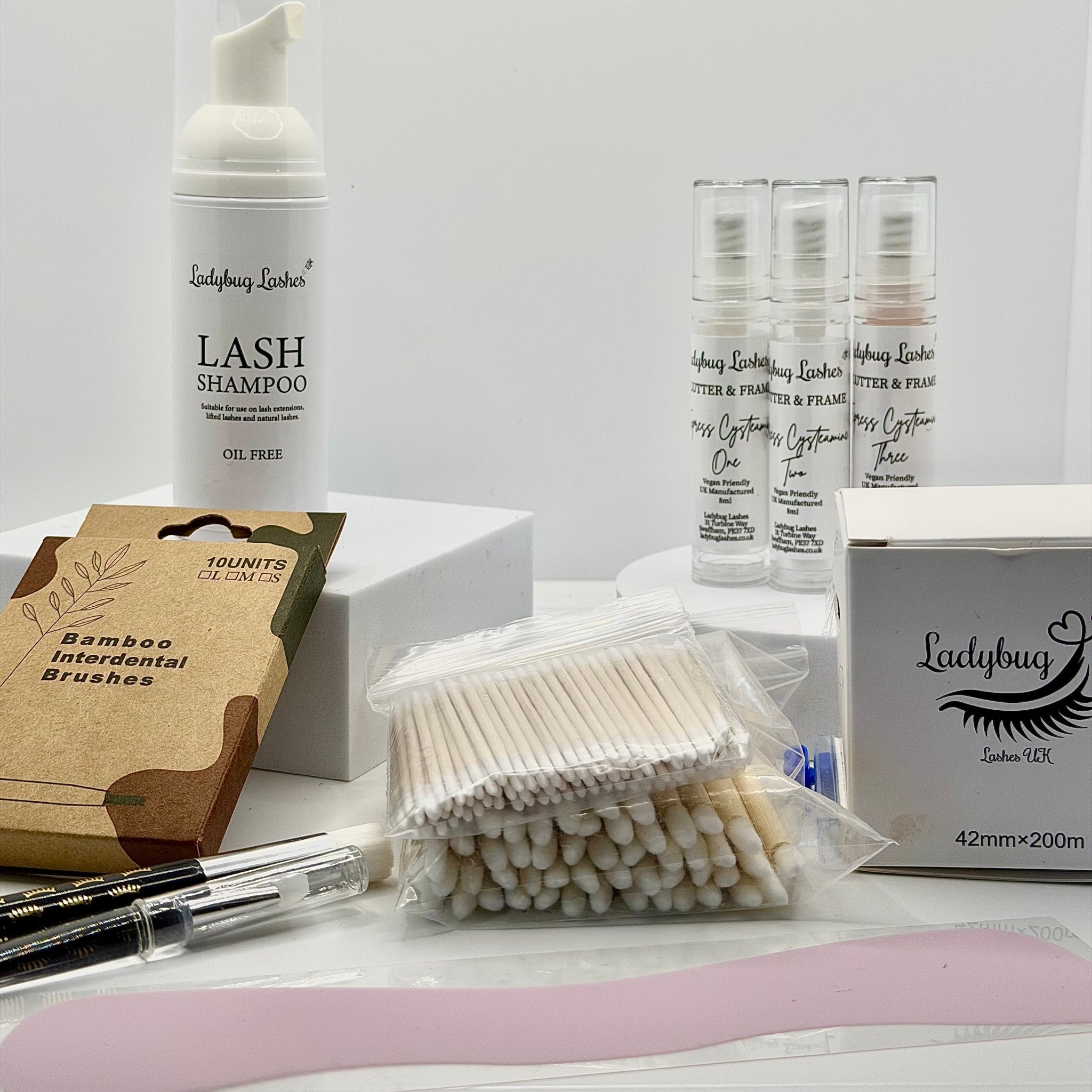 Ladybug Lashes Express Cysteamine Brow Lamination Kit flatlay showing Ladybug Lashes Lash Shampoo foam pump, three Flutter and Frame Express Cysteamine airless pump bottles One Two and Three, Ladybug Lashes 42mm x 200m brow lamination wrap box, bamboo interdental brushes pack, cotton buds, microfibre brushes and pink silicone spatula on white background
