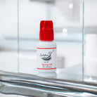A bottle of Spring UK adhesive/glue with a red cap on a reflective surface, branded by Ladybug Lashes.