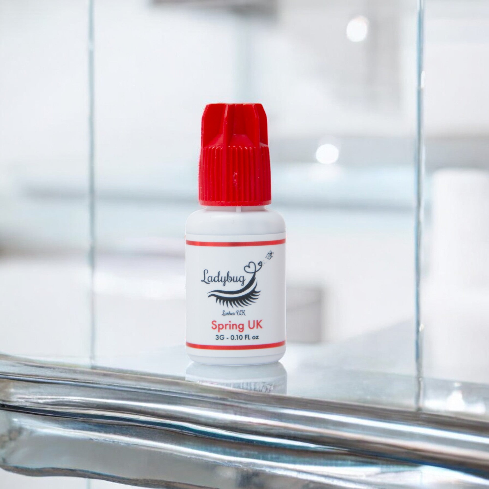A bottle of Spring UK adhesive/glue with a red cap on a reflective surface, branded by Ladybug Lashes.