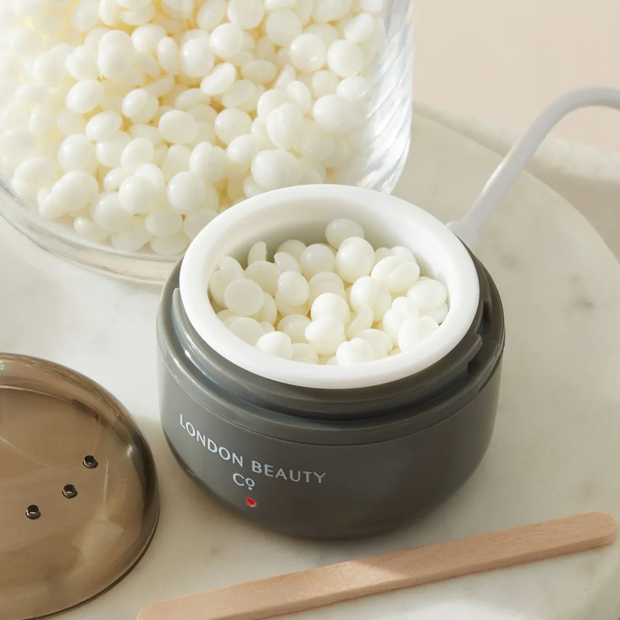 London Brow Flexi Mini Wax Refill Bags lifestyle shot showing white pearl-shaped wax beads loaded into an open dark grey London Beauty Co wax heater pot with a wooden spatula and glass bowl of wax beads in background on marble surface 
