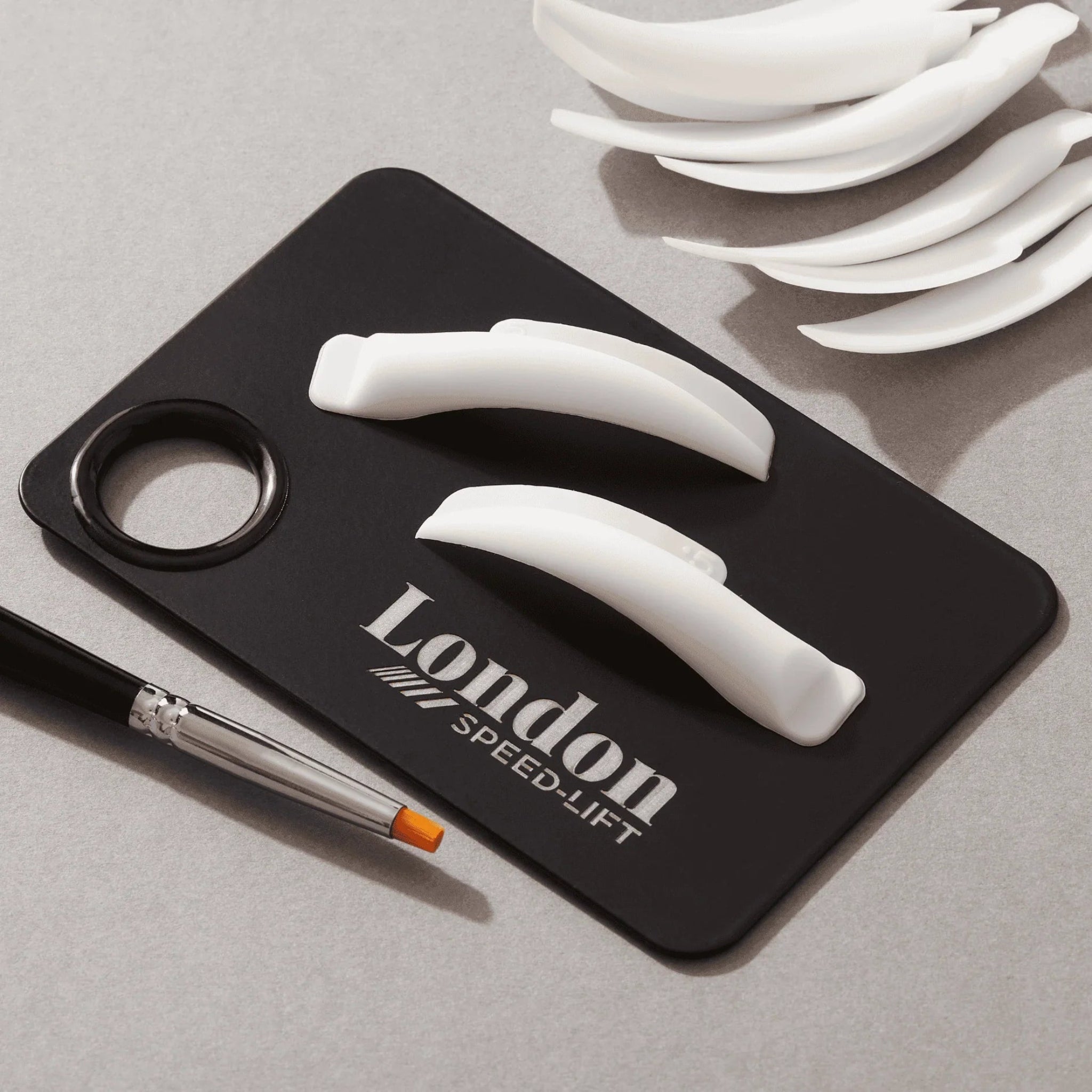London Brow Company Speed-Lift Lash Lift and Tinting Course tool detail shot showing two white curved silicone lash lift shields placed on a London Speed-Lift branded black silicone work mat with thumb hole alongside a flat angled brush and a stack of additional white silicone shields from diagonal angle on grey background