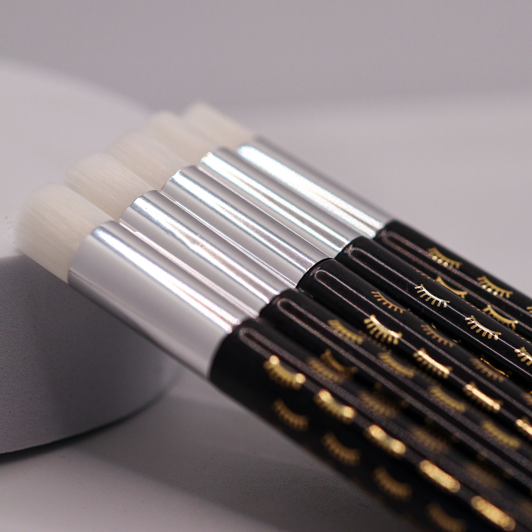 Ladybug Lashes Soft Bristle Lash Cleansing Brushes pack of 5 product shot showing five brushes fanned out in a row with white dome-shaped soft bristle heads silver ferrules and black handles printed with gold and white closed-eye lash motifs from diagonal overhead angle on white background