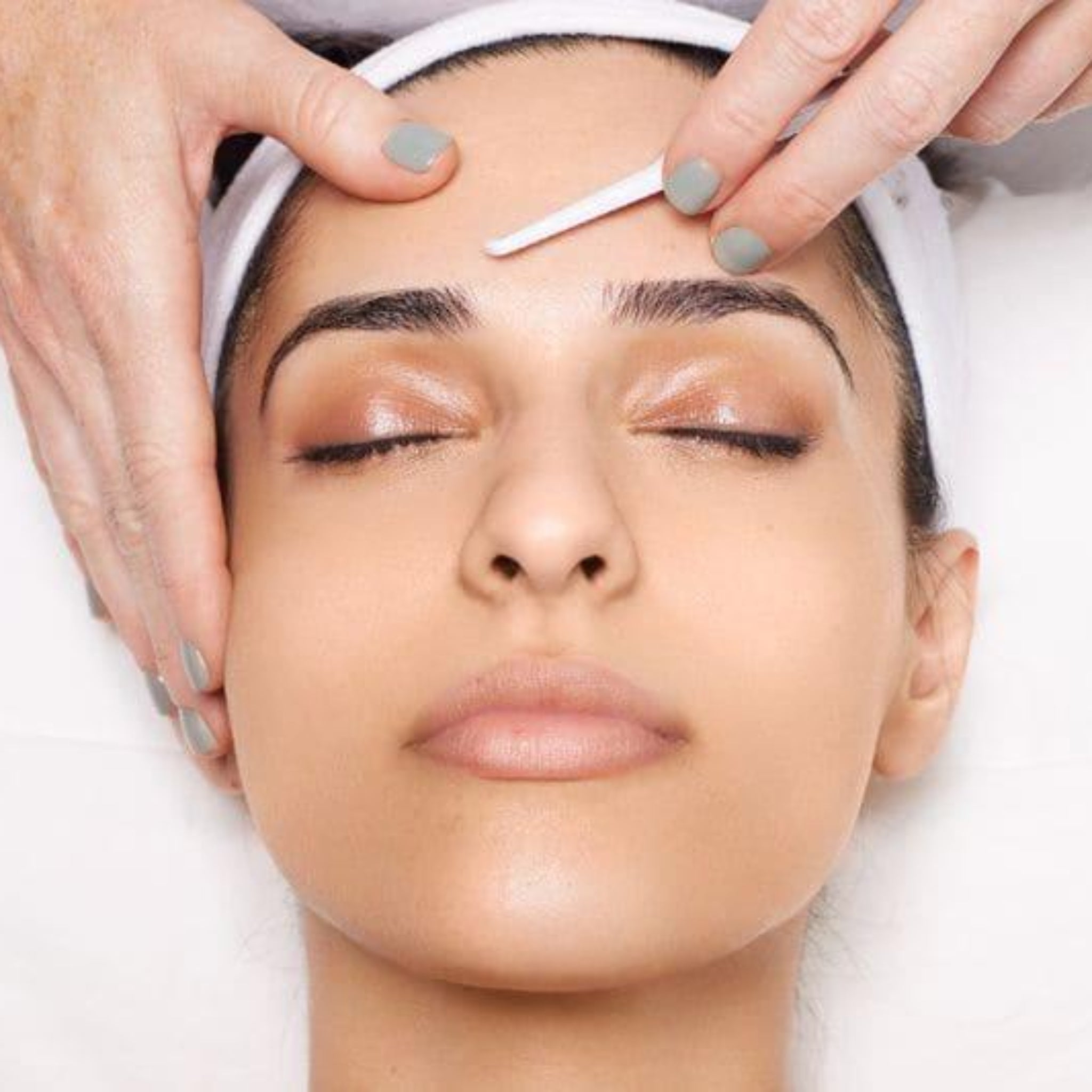 London Brow Company Glass Skin Dermaplaning Facials Online Course treatment shot showing therapist performing dermaplaning on a female client's forehead using a small blade tool with client lying reclined eyes closed wearing a white headband on white treatment bed
