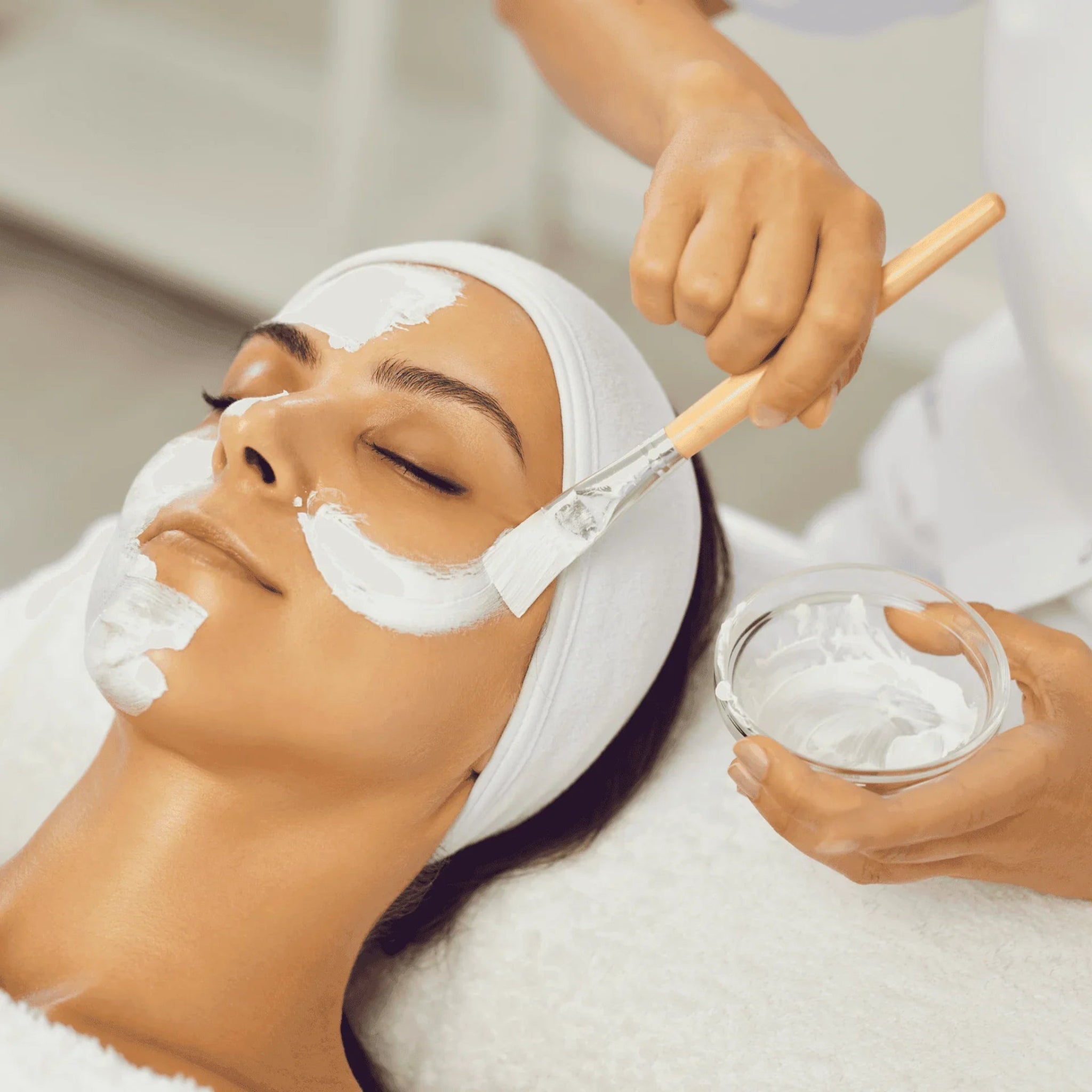 London Brow Company Facials and Facial Therapy Online Course treatment shot showing therapist applying white cream face mask to a reclined female client using a wooden-handled brush with glass bowl of product in hand client lying eyes closed wearing white headband on white treatment bed