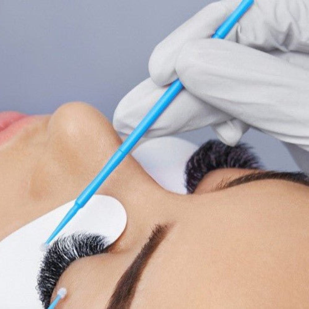 Ladybug Lashes Classic Lash Online Training Course in-treatment close-up showing white-gloved lash technician applying individual classic lash extensions with a blue micro applicator brush to a fair-skinned female client with eyes closed and white under-eye pad from diagonal overhead angle on grey background