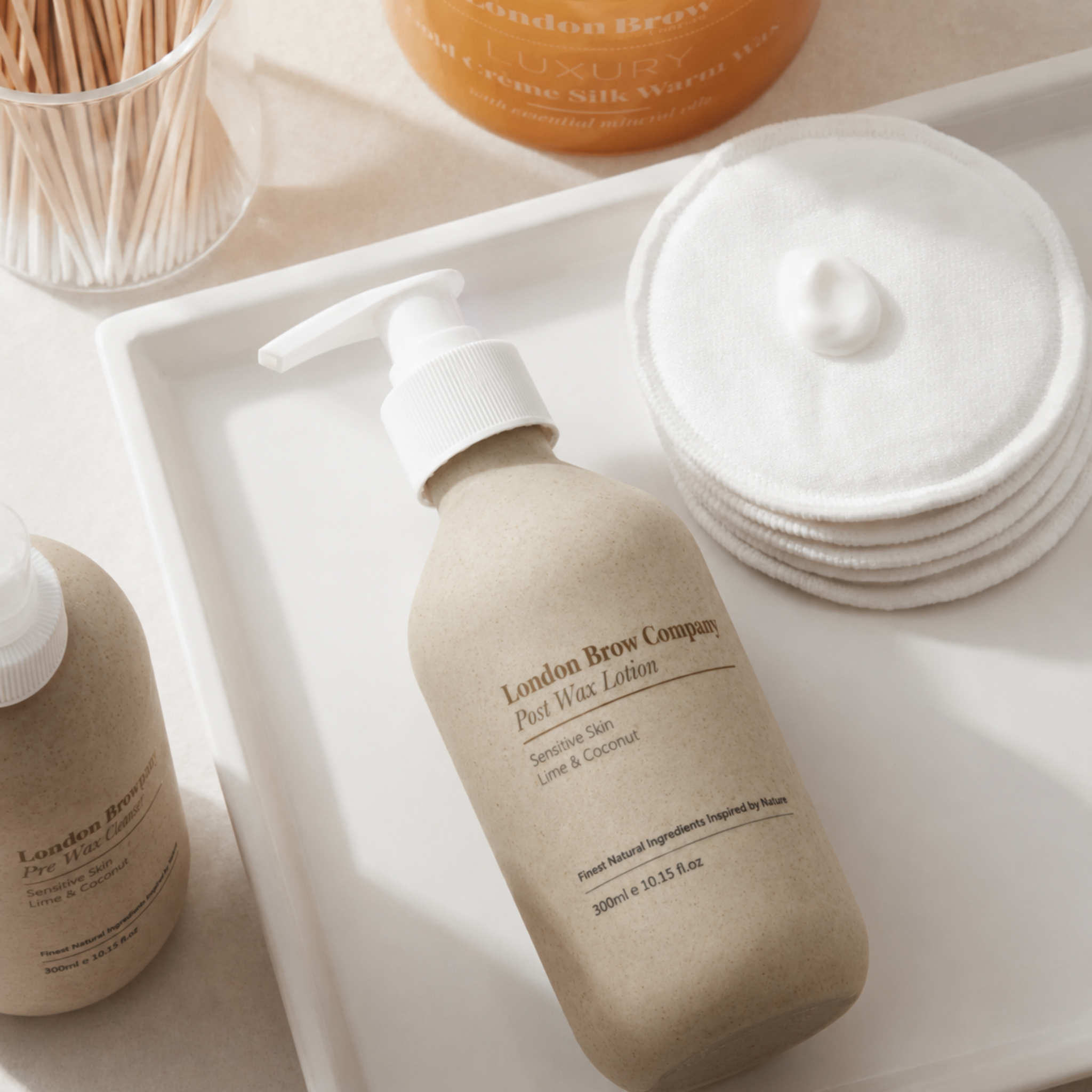 The London Brow Company Post-Wax Lotion lifestyle product shot showing 300ml stone-finish pump bottle labelled Post Wax Lotion Sensitive Skin Lime & Coconut on white tray alongside Pre Wax Cleanser bottle cotton pads and wooden cotton buds from diagonal overhead angle on cream background