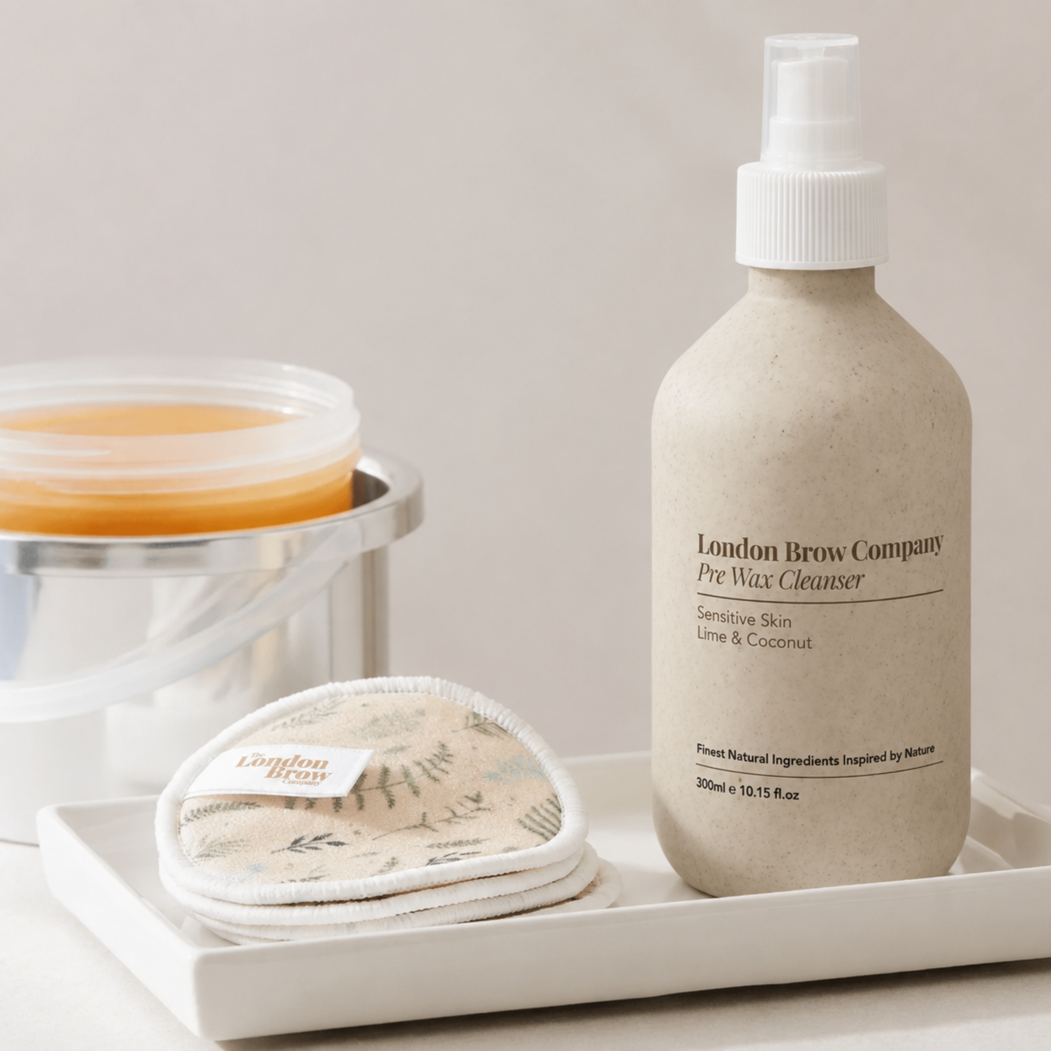 The London Brow Company Pre-Wax Cleanser lifestyle product shot showing 300ml stone-finish pump bottle labelled Pre Wax Cleanser Sensitive Skin Lime & Coconut on white tray alongside London Brow Company branded reusable cotton pads and wax heater with amber wax from diagonal angle on cream background