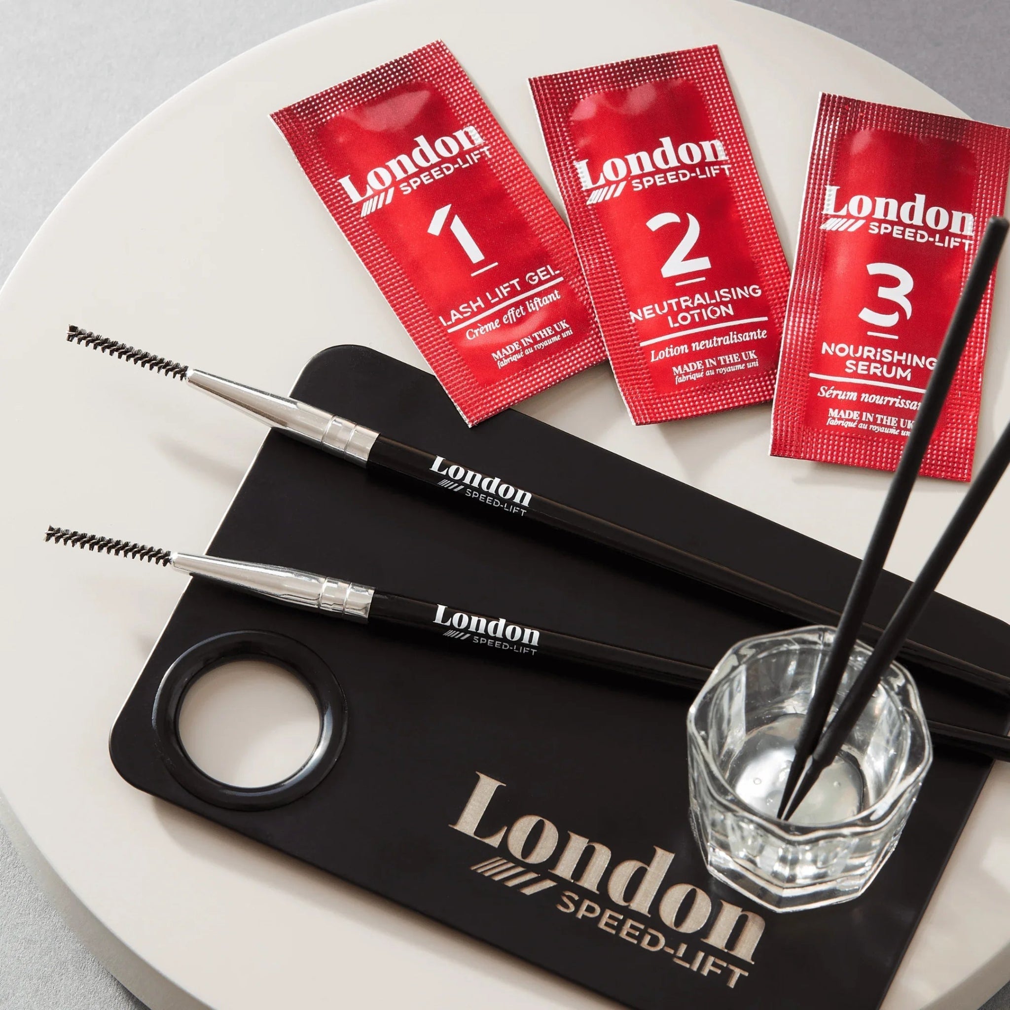 London Brow Company Speed-Lift Lash Lift and Tinting Course tool detail shot showing two white curved silicone lash lift shields placed on a London Speed-Lift branded black silicone work mat with thumb hole alongside a flat angled brush and a stack of additional white silicone shields from diagonal angle on grey background