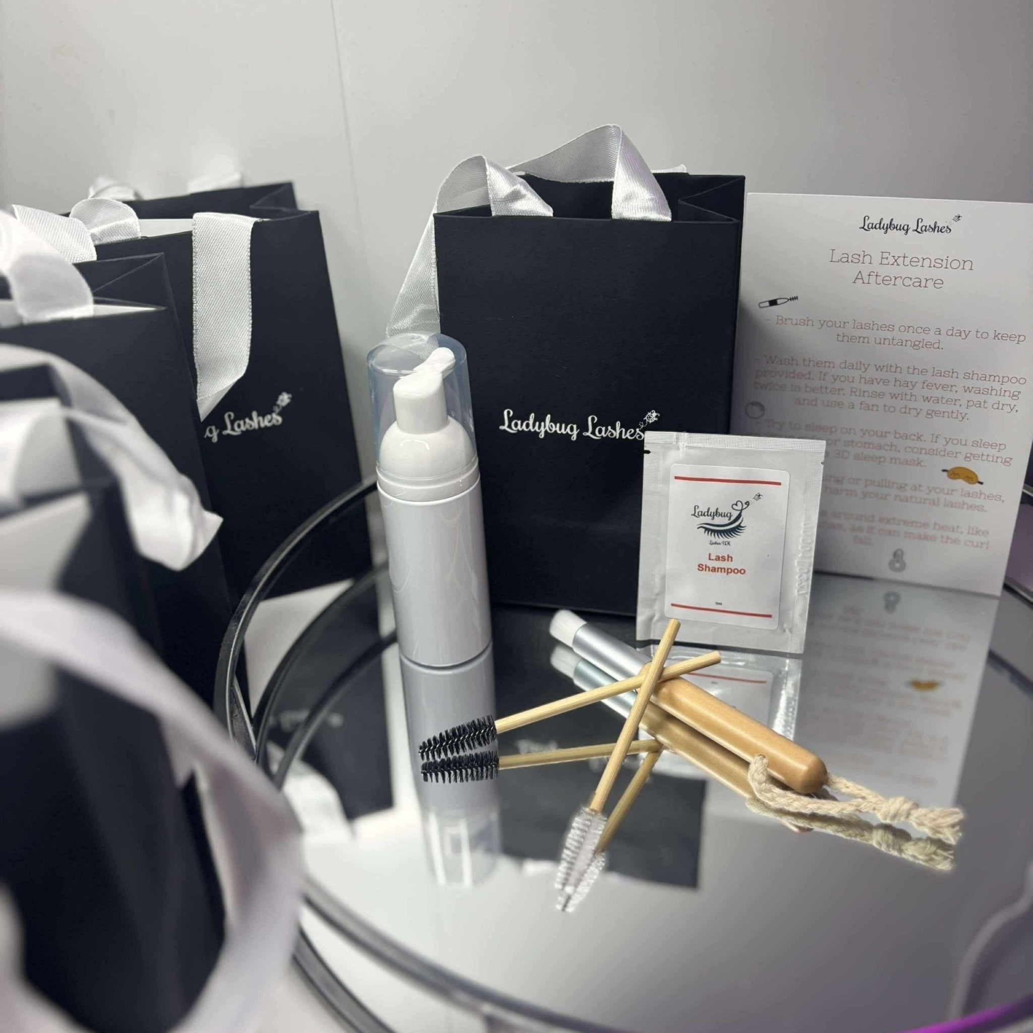 Ladybug Lashes Lash Shampoo Sachet Kit contents displayed on a reflective glass surface surrounded by black Ladybug Lashes gift bags with white satin handles, including a white foam pump bottle, a lash shampoo sachet, a bamboo spoolie, a bamboo cleansing brush with macramé loop, a mini applicator, and a printed Lash Extension Aftercare card