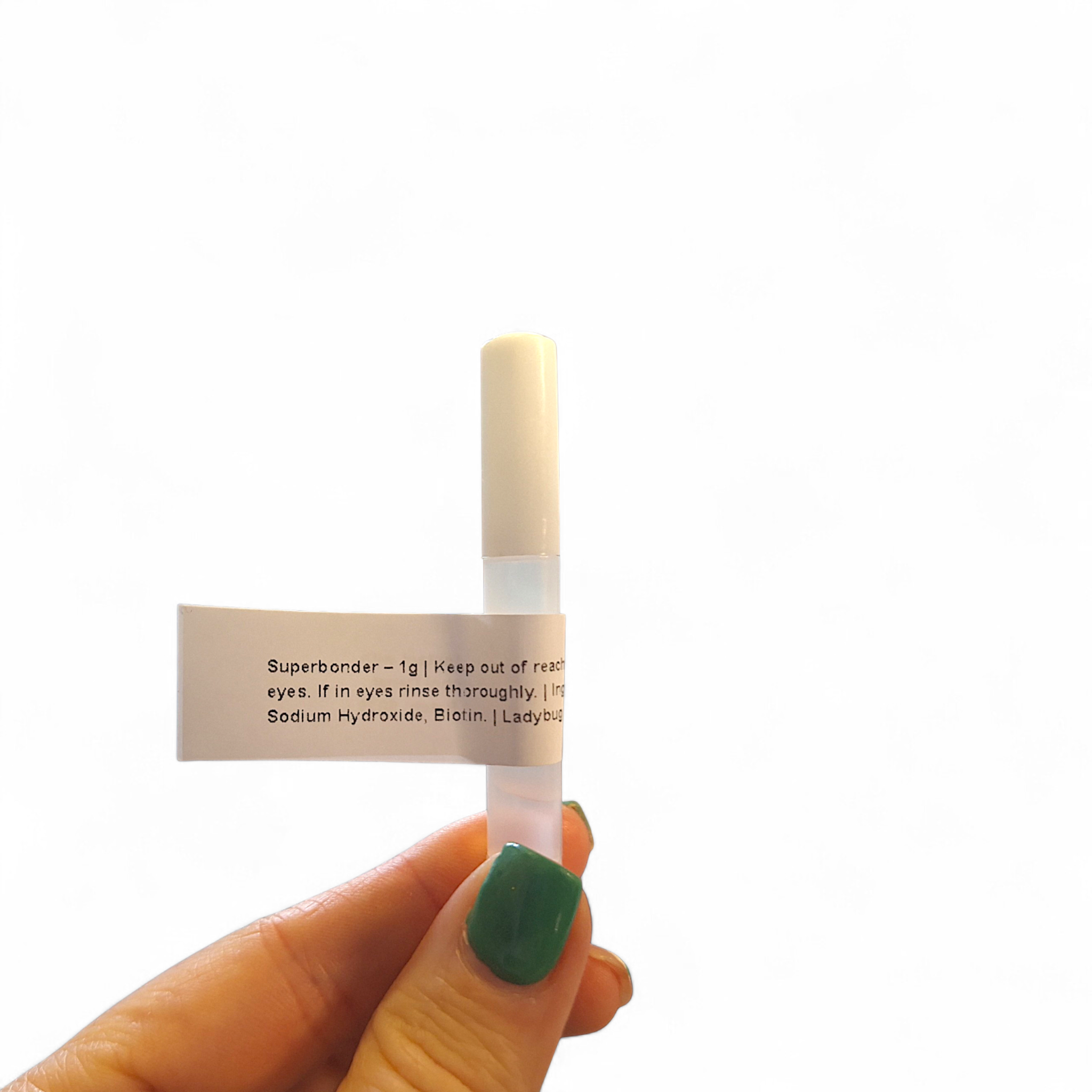 Hand with green nail polish holding a single Ladybug Lashes SuperBonder 1g sample bottle against a white background, showing the white label with "Superbonder – 1g" text and partial ingredients list including Sodium Hydroxide and Biotin with Ladybug Lashes branding