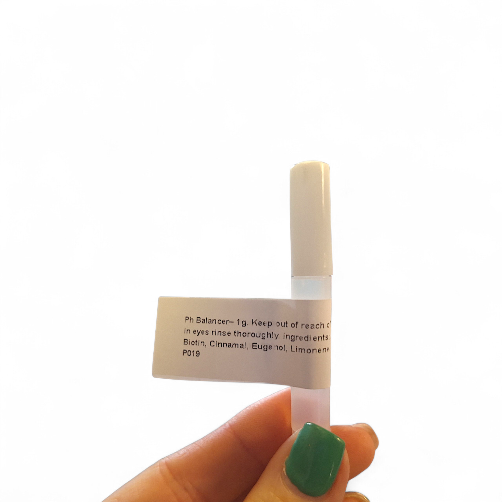 Hand with green nail polish holding a single Ladybug Lashes PH Balancer 1g sample bottle against a white background, showing the white label with "Ph Balancer – 1g" text and partial ingredients list including Biotin, Cinnamal, Eugenol and Limonene
