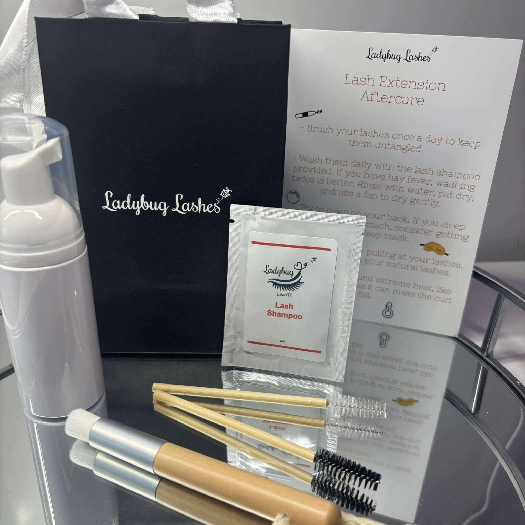 Ladybug Lashes Lash Shampoo Sachet Kit contents laid out on a glass surface in front of a black Ladybug Lashes gift bag with white satin handles, including a white foam pump bottle, a Ladybug Lashes lash shampoo sachet, a bamboo spoolie brush, a bamboo cleansing brush with macramé loop, a mini spray bottle, and a printed Lash Extension Aftercare instruction card