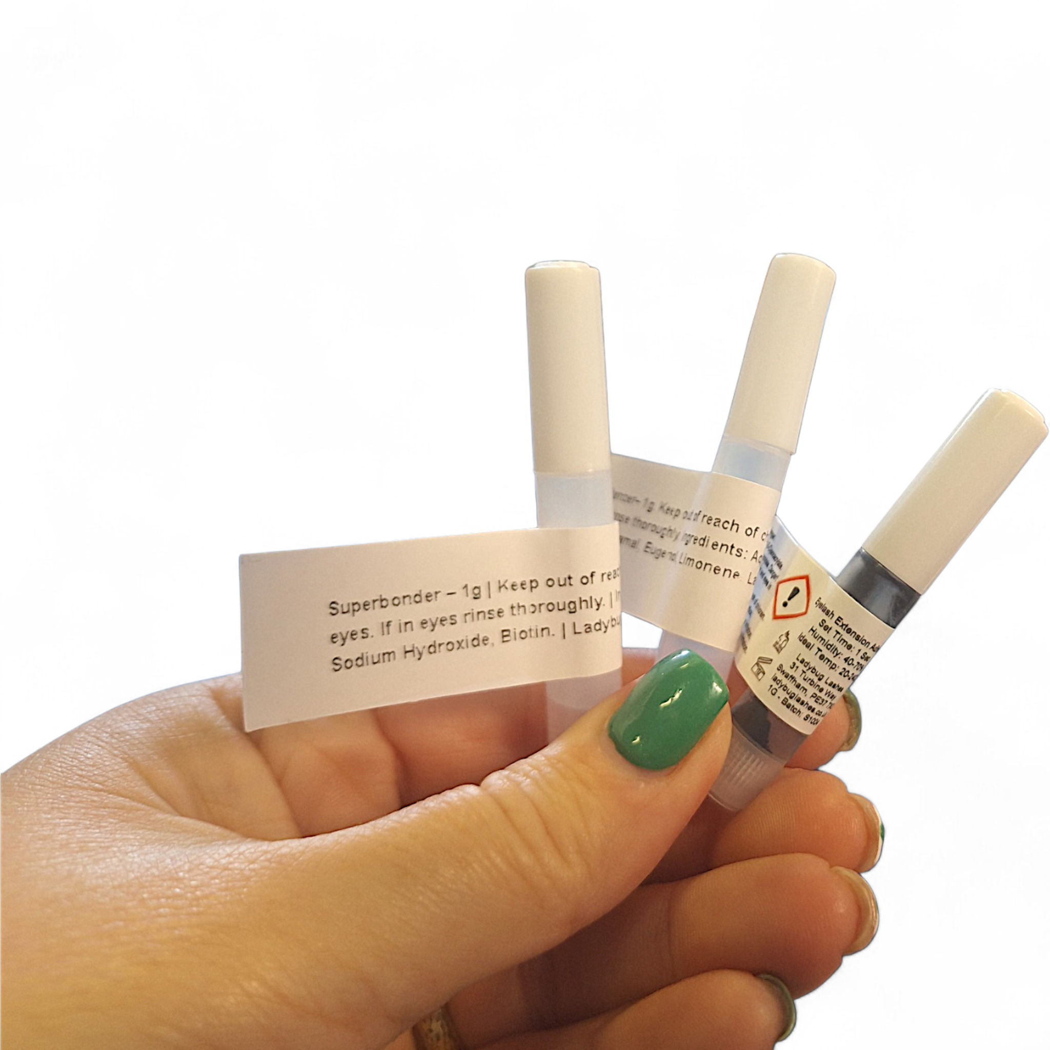 Hand with green nail polish holding three Ladybug Lashes 1g sample bottles fanned out against a white background — a SuperBonder sample with white label showing ingredients and safety information, a clear liquid sample bottle, and a lash extension adhesive sample bottle with black cap and hazard warning label visible