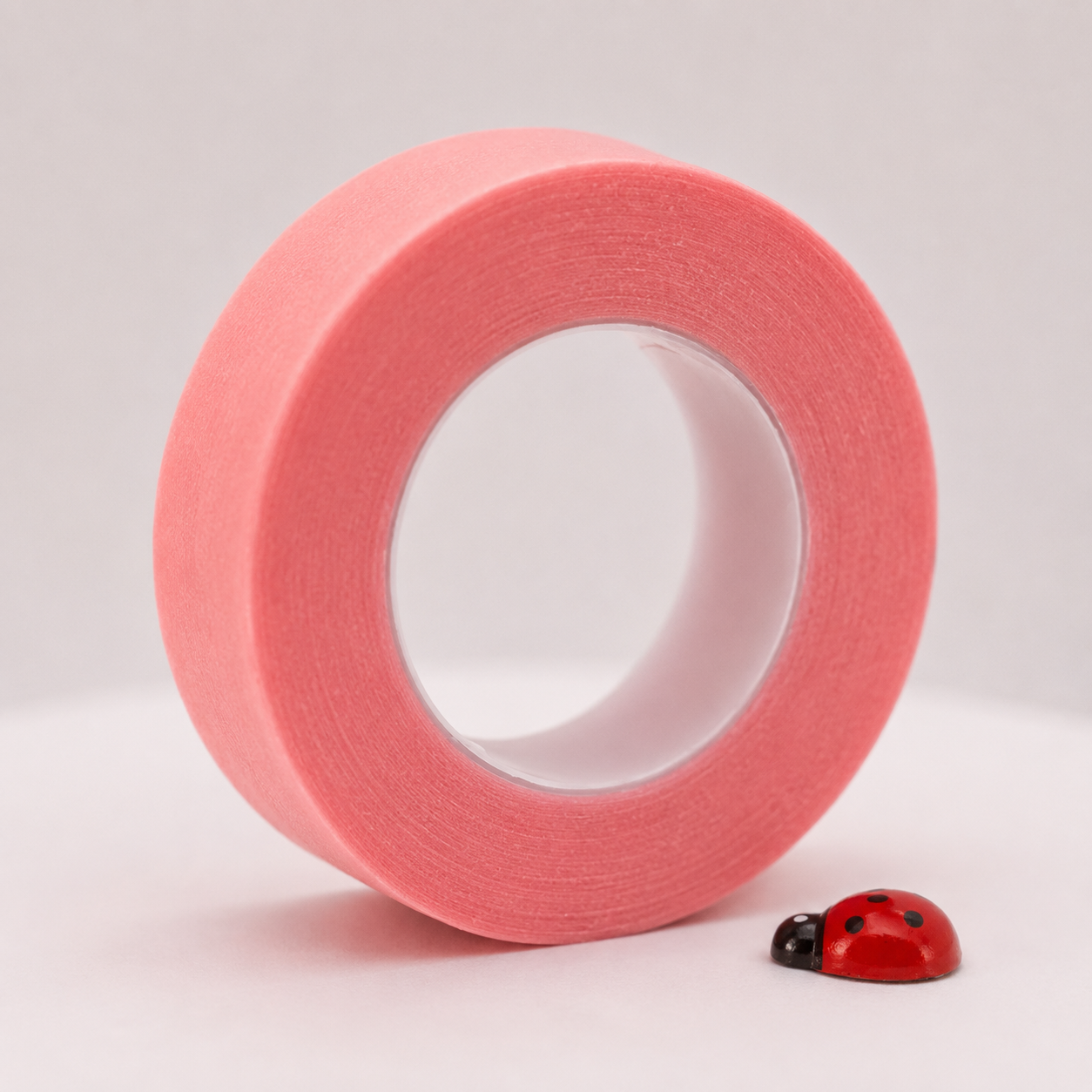 Single large roll of Ladybug Lashes Gentle Silicone Eye Tape in pale pink standing upright against a light grey background, with a small red and black ladybird ornament beside it — showing the smooth silicone texture and wide format of the tape used for securing lower lashes during lash extension treatments