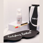 Ladybug Lashes Lash Shampoo Kit with Eye Mask contents showing a black Ladybug Lashes branded sleep mask in the foreground, a Ladybug Lashes Bubblegum Shampoo foam pump bottle and bamboo-handled lash cleansing brush with red spoolie on a white plinth, and a black Ladybug Lashes gift bag with white ribbon handles on a white surface