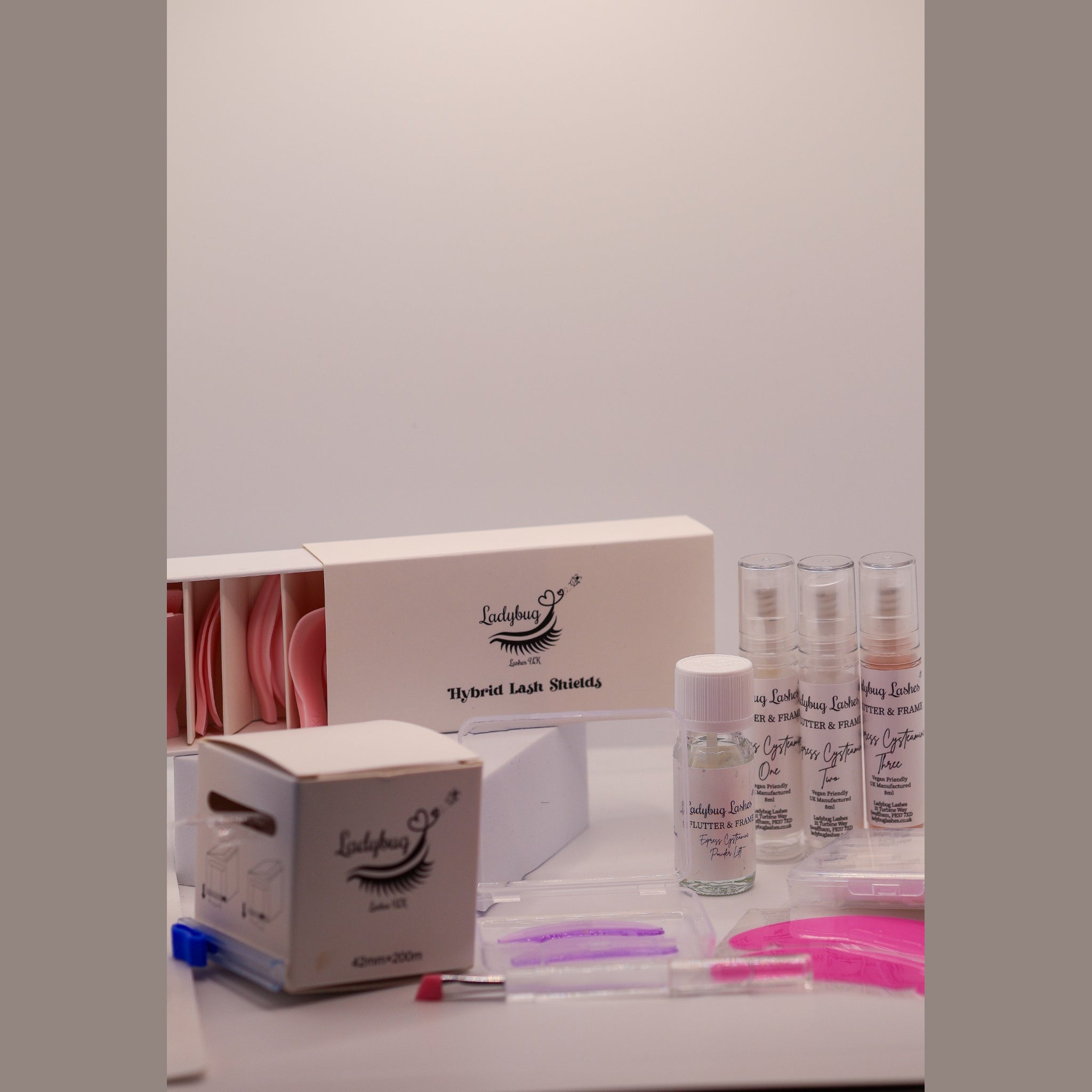 Ladybug Lashes Korean-Inspired Lash Lift Masterclass kit contents showing open Ladybug Lashes Hybrid Lash Shields box with pink silicone shields, Ladybug Lashes cling film dispenser box, three Ladybug Lashes Flutter and Frame Express Cysteamine airless pump bottles, Powder Lift bottle, coloured 