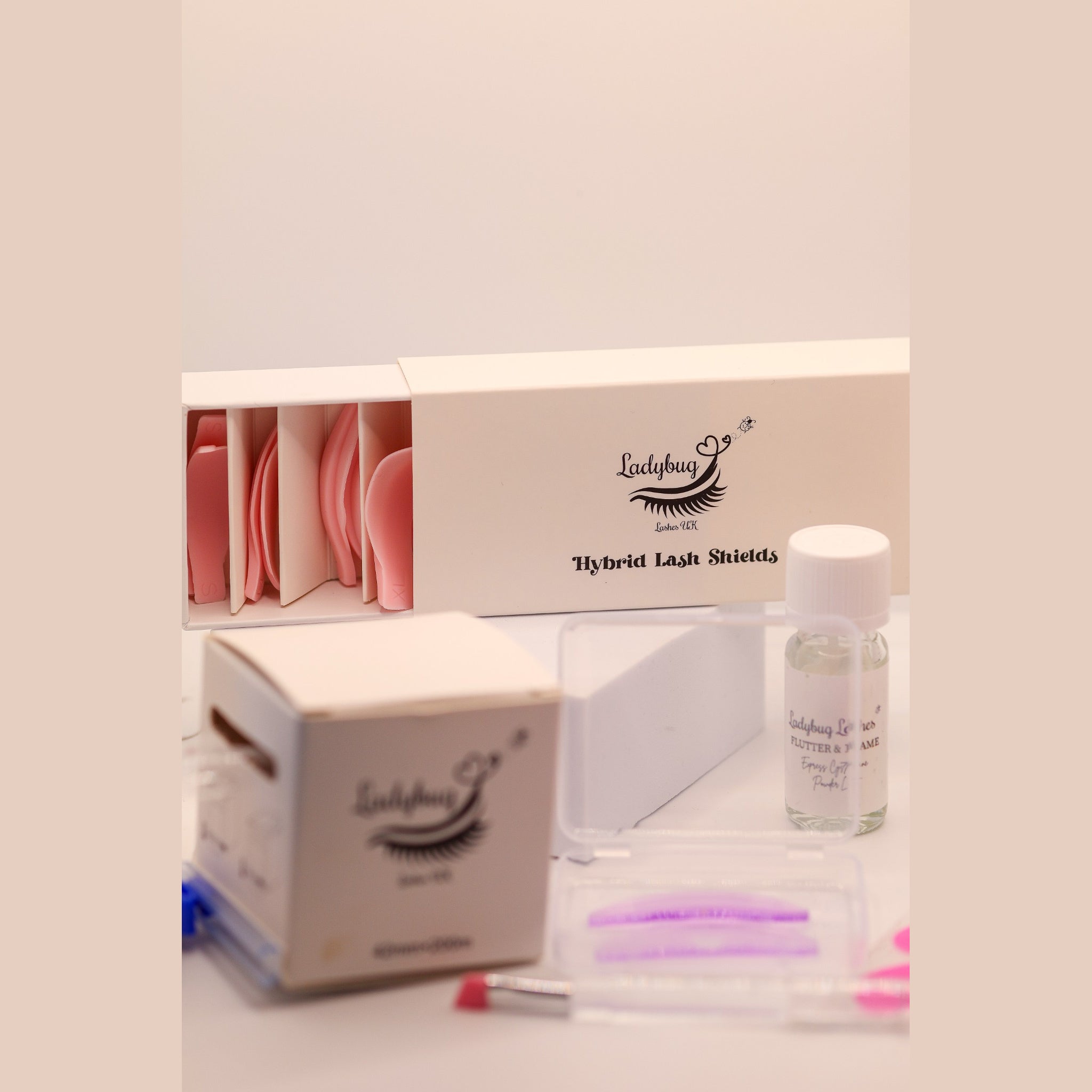 Close-up of Ladybug Lashes Flutter and Frame Express Cysteamine One Two Three airless pump bottles and Powder Lift bottle alongside Ladybug Lashes Hybrid Lash Shields box with pink and purple silicone lash shields in the foreground on a white surface with pink border