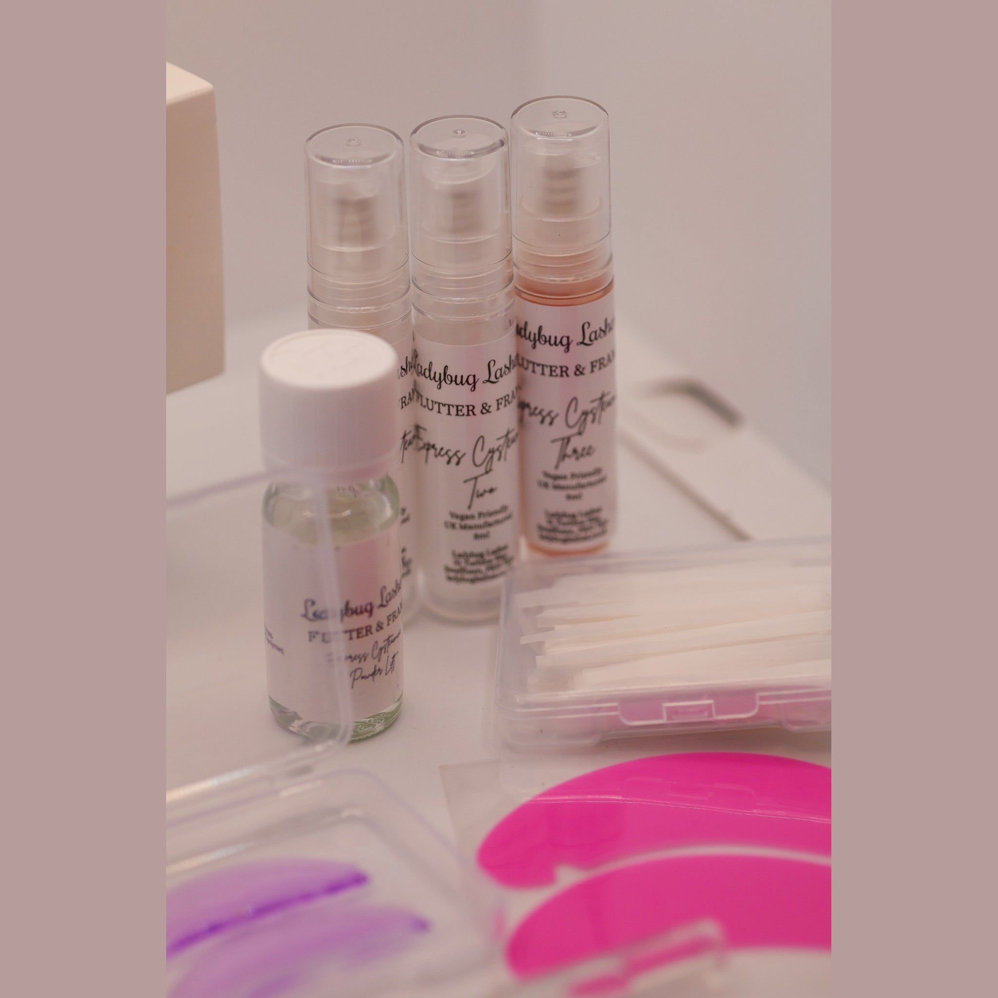 Close-up of Ladybug Lashes Flutter and Frame Express Cysteamine Two and Three airless pump bottles and Powder Lift bottle with pink and purple silicone lash shields in the foreground and cotton buds and a clear storage case in the background on a white surface with pink border