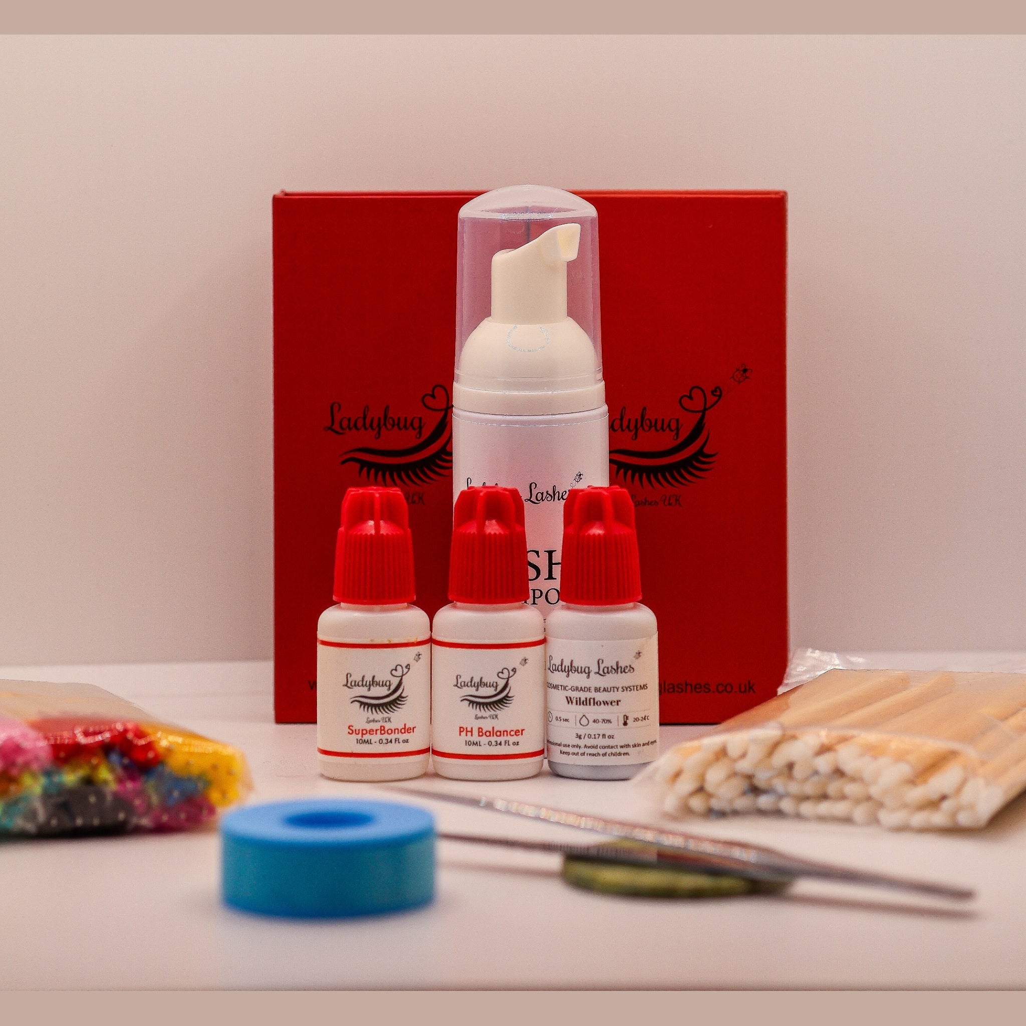 Ladybug Lashes Beginner Lash Starter Kit flatlay showing a white foam lash shampoo pump bottle, three white and red Ladybug Lashes bottles labelled SuperBonder, pH Balancer and Wildflower adhesive, blue gel tape roll, bamboo applicator wands, coloured mascara spoolies and tweezers, arranged in front of a red Ladybug Lashes branded gift box on a white surface