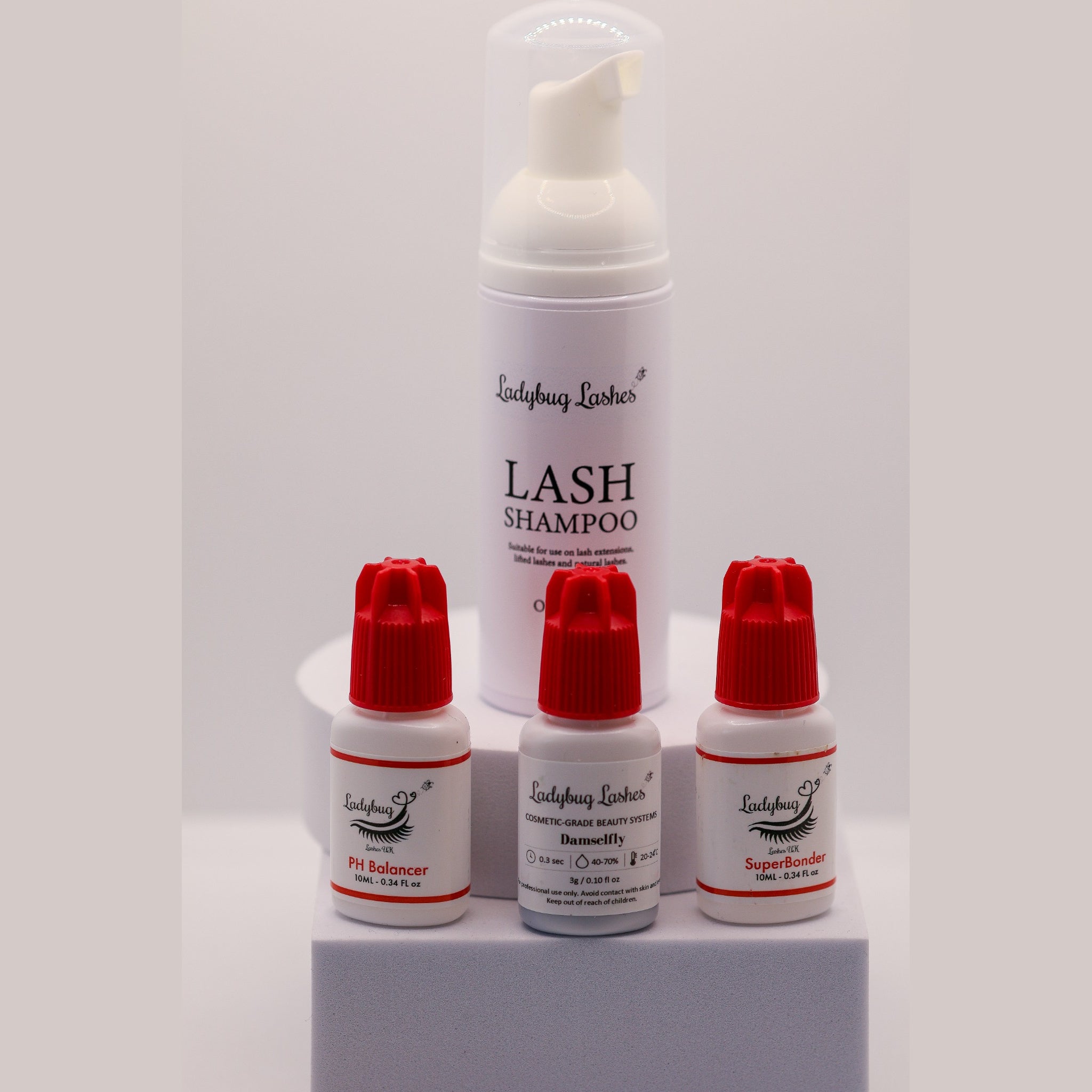 Ladybug Lashes Retention Trio showing four products on a white background: Lash Shampoo 25ml foam pump bottle, Booster retention booster 10ml bottle with red cap, PH Balancer 10ml bottle with red cap, and Meadow adhesive 1g bottle with red cap
