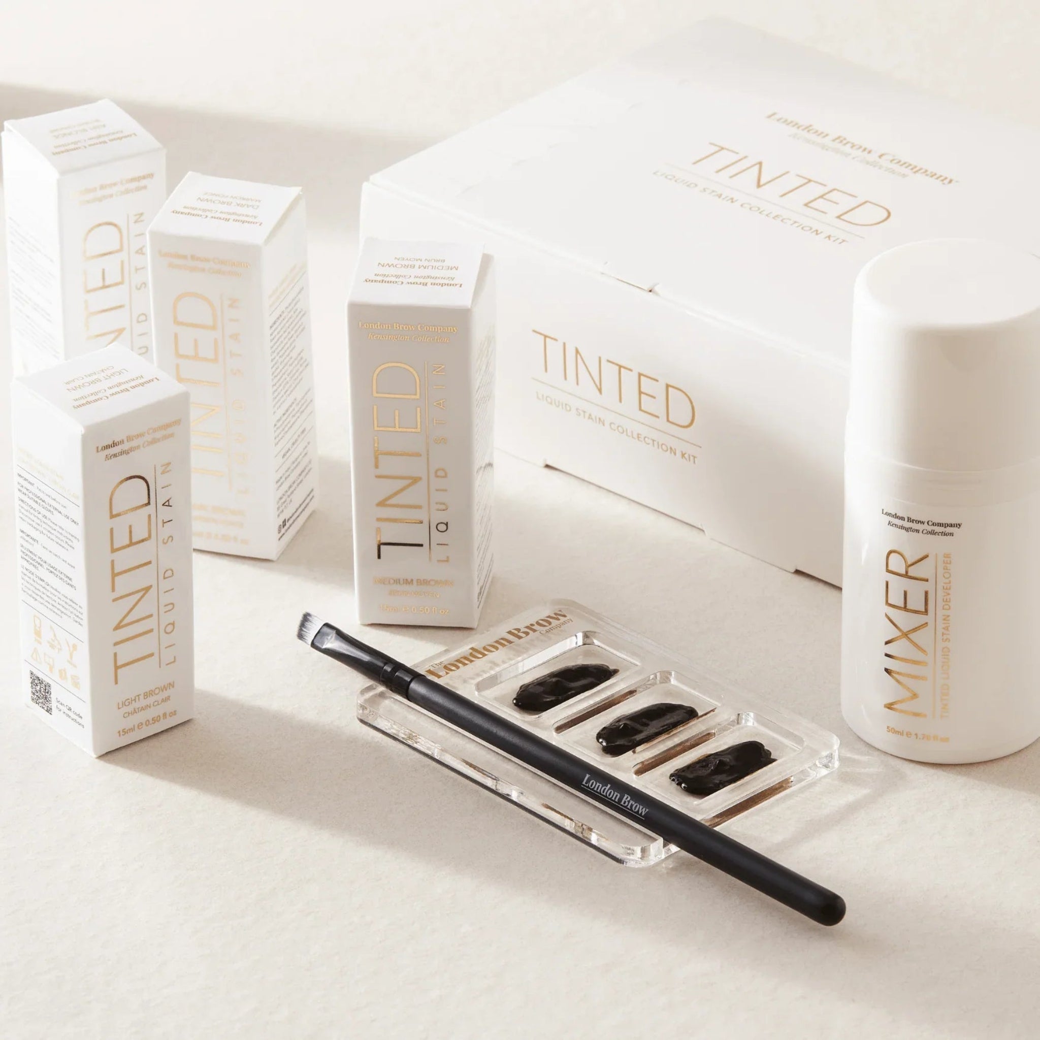 London Brow Company Kensington Collection Tinted Liquid Brow Stain 15ml lifestyle flatlay showing multiple shade bottles and retail boxes arranged on a warm cream surface from diagonal overhead angle